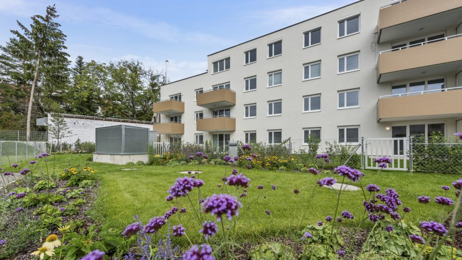 Mehrfamilienhaus mit Garten und Blumen im Vordergrund an einem sonnigen Tag.