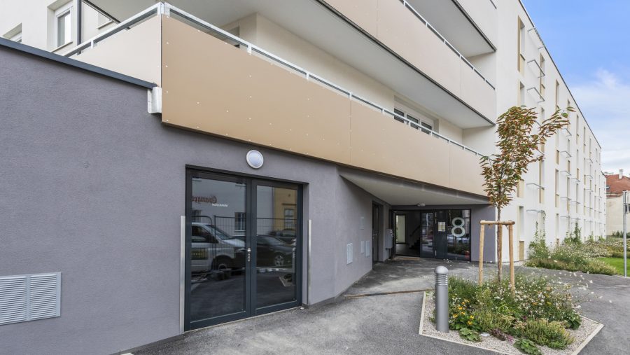 Moderne Hausfassade mit Balkon und Eingangstür, umgeben von kleinen Gartenflächen und einem gepflanzten Baum.