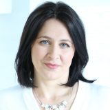 Eine Frau mit dunklen Haaren und einer Halskette in einem weißen Oberteil lächelt leicht in die Kamera.