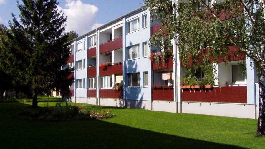 Mehrfamilienhaus mit Balkonen und grüner Rasenfläche im Vordergrund, umgeben von Bäumen und einem blauen Himmel.