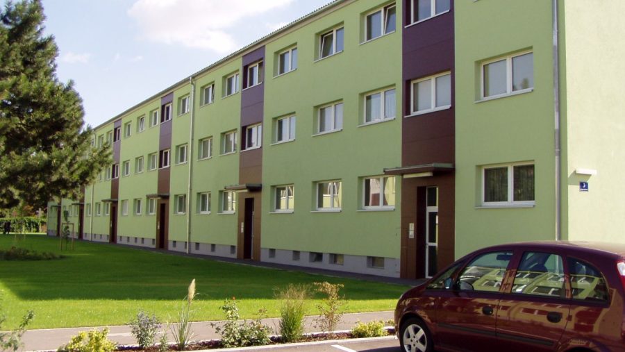 Grünes Wohnhaus mit mehreren Etagen, umgeben von Rasenfläche und geparktem Auto im Vordergrund.