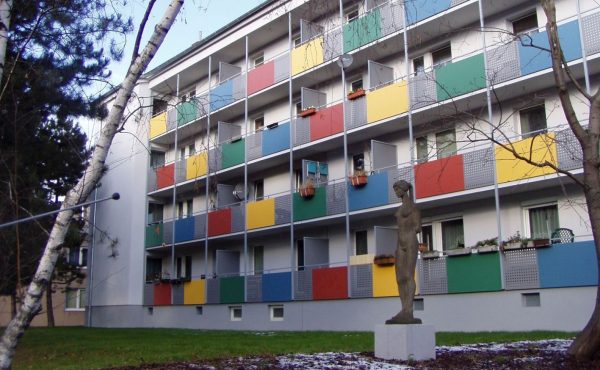 Mehrstöckiges Wohngebäude mit bunten Balkonen und einer Statue im Vordergrund, umgeben von Bäumen.