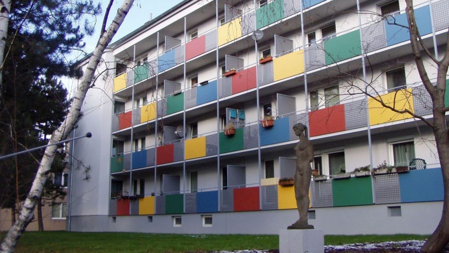 Mehrstöckiges Wohngebäude mit bunten Balkonen und einer Statue im Vordergrund, umgeben von Bäumen.