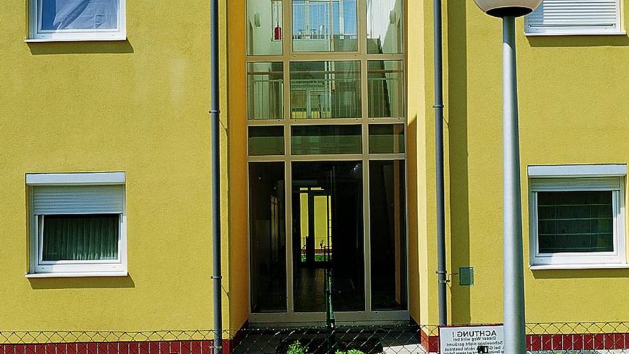 Gelbes Gebäude mit Glasfassade und hohen Fenstern, im Vordergrund ein Zaun und eine moderne Straßenlaterne.