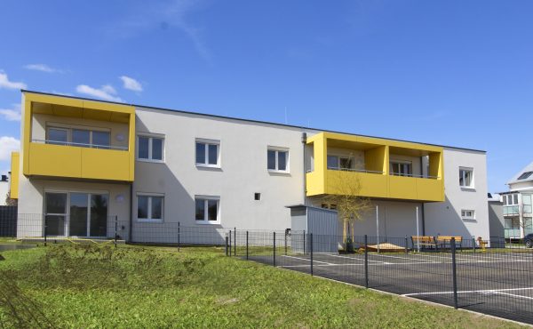 Modernes zweistöckiges Gebäude mit gelben Balkonen und umzäuntem Vorgarten bei blauem Himmel.