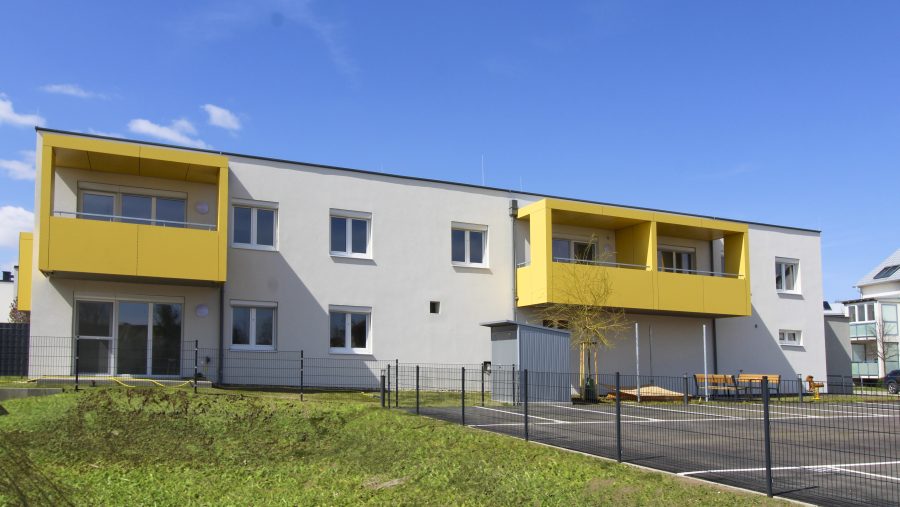 Modernes zweistöckiges Gebäude mit gelben Balkonen und umzäuntem Vorgarten bei blauem Himmel.