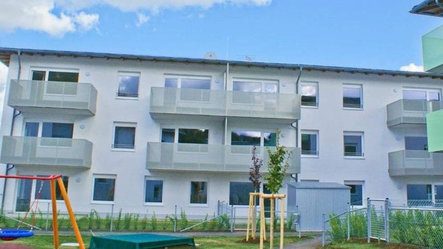 Mehrfamilienhaus mit Balkonen an einem sonnigen Tag, umgeben von Rasen und einem Spielplatz im Vordergrund.