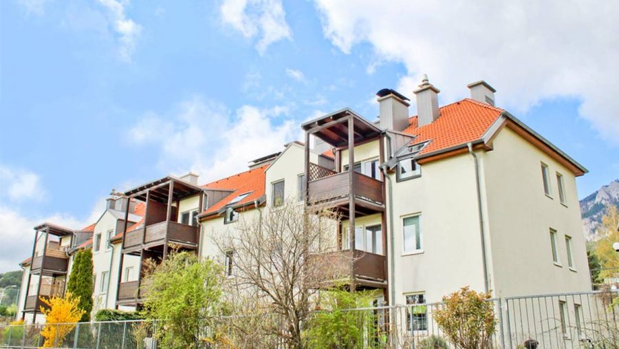 Mehrfamilienhaus mit roten Dächern, Balkonen und bewölktem Himmel im Hintergrund.