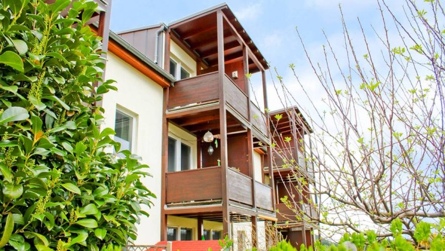 Mehrstöckiges Gebäude mit Holzveranda und grünen Pflanzen im Vordergrund.