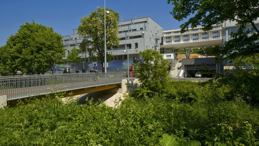 Gebäude mit moderner Architektur hinter Brücke und grüner Landschaft bei sonnigem Wetter.