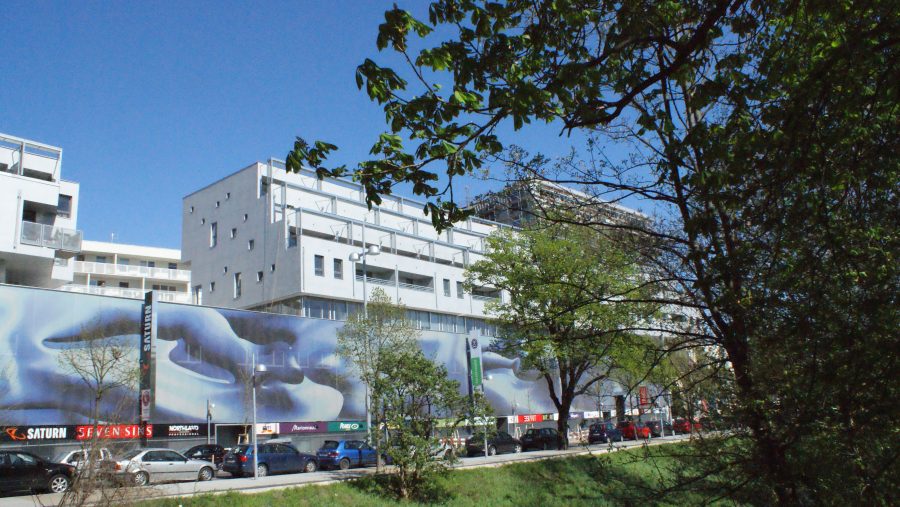 Straßenansicht mit modernen Gebäuden im Hintergrund und grüner Vegetation im Vordergrund bei klarem Himmel.