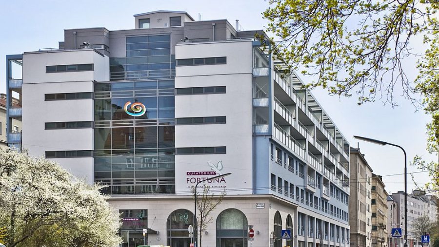 Modernes Bürogebäude mit Glasfassade in städtischer Umgebung, Frühling, blühende Bäume im Vordergrund.