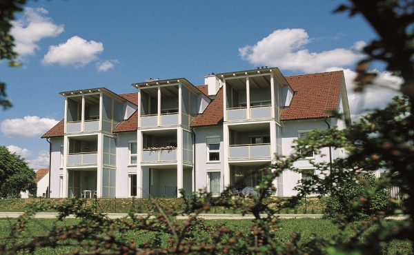 Modernes Mehrfamilienhaus mit Balkonen unter blauem Himmel und Wolken, umgeben von grüner Vegetation.