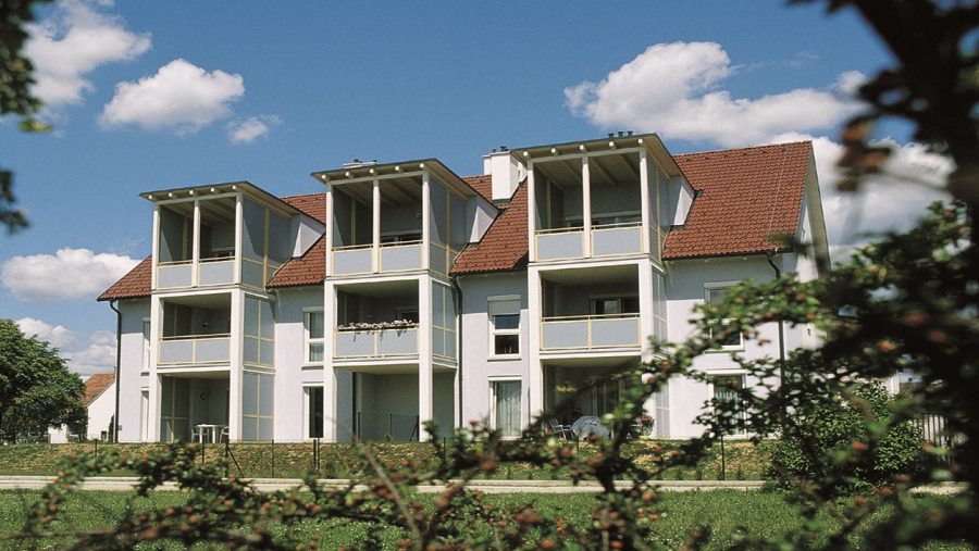 Modernes Mehrfamilienhaus mit Balkonen unter blauem Himmel und Wolken, umgeben von grüner Vegetation.