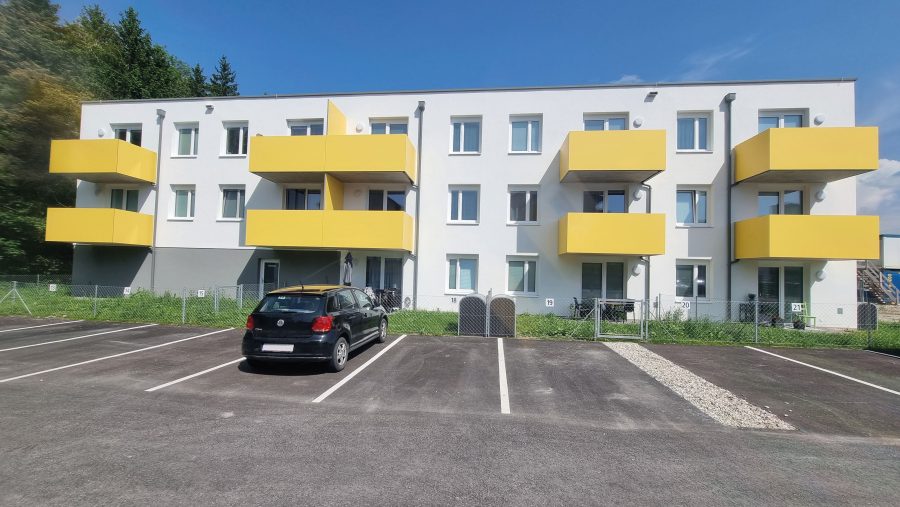 Mehrfamilienhaus mit gelben Balkonen, Parkplatz im Vordergrund, blauer Himmel im Hintergrund.
