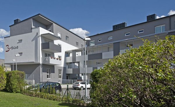 Mehrfamilienhaus mit Balkonen und geparkten Autos, sonniger Tag, blauer Himmel, umgeben von grüner Vegetation.