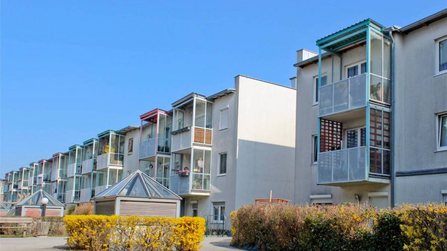 Mehrfamilienhäuser mit Glasbalkonen und blauem Himmel im Hintergrund.