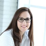 Frau mit Brille lächelt in Kamera, modernes Bürohintergrund.