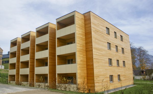 Modernes Wohngebäude aus Holz mit Balkonen und Naturumgebung unter blauem Himmel.