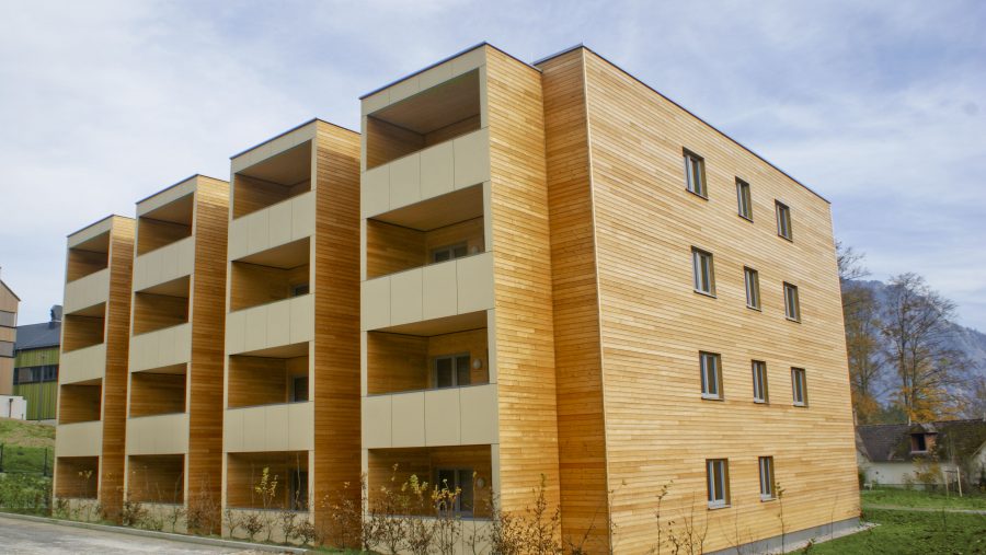Modernes Wohngebäude aus Holz mit Balkonen und Naturumgebung unter blauem Himmel.
