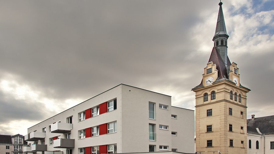 Moderne Wohnarchitektur neben historischer Kirchturm. Wolkiger Himmel im Hintergrund.