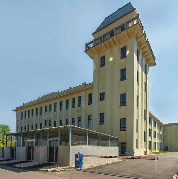 Revitalisierung „Alte Textilspinnerei“ in Teesdorf/NÖ Großes, historisches Gebäude mit Turm, umgeben von blauem Himmel und Vegetation, bei Tageslicht gesehen.