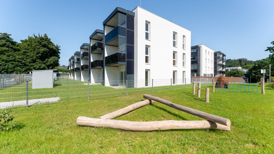 Moderne Wohngebäude mit Balkonen und eingezäunten Grünflächen bei sonnigem Wetter.