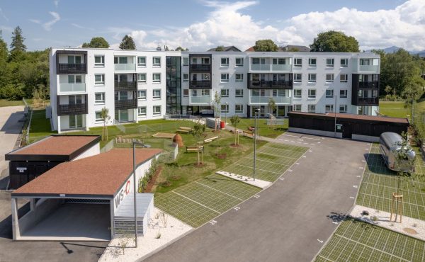 Mehrstöckiges Wohngebäude mit grünem Hof und Parkplatz bei sonnigem Wetter.