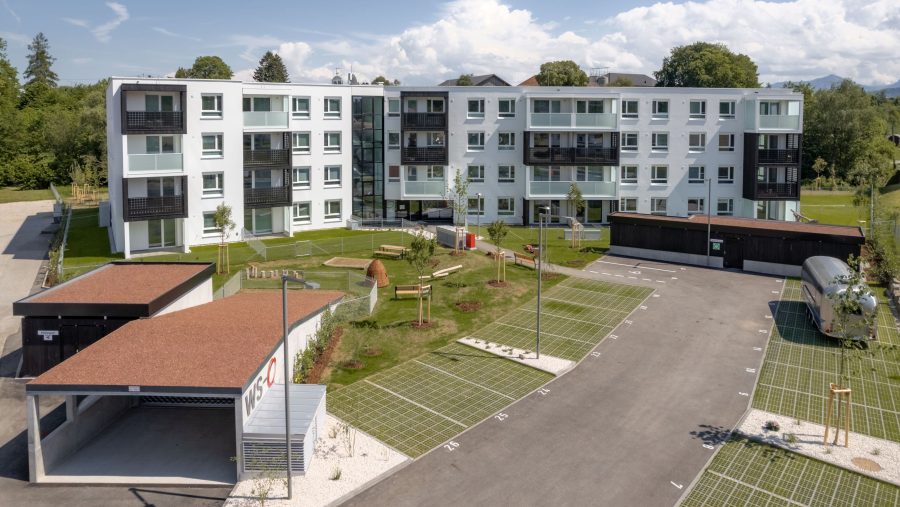 Mehrstöckiges Wohngebäude mit grünem Hof und Parkplatz bei sonnigem Wetter.