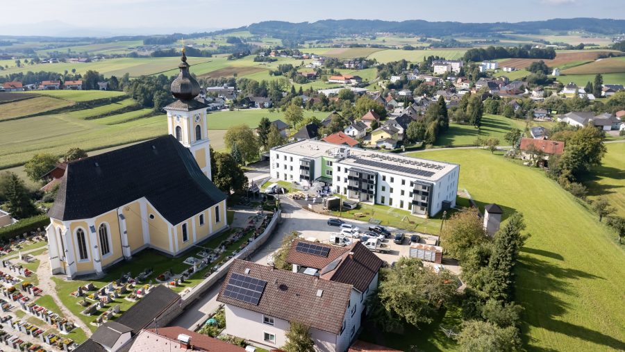 Luftaufnahme eines Dorfes mit Kirche, modernen Gebäuden und grüner Landschaft im Hintergrund.