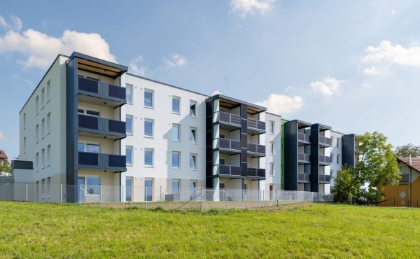 Mehrstöckiges Wohngebäude mit Balkon und Grünfläche im Vordergrund bei sonnigem Himmel.