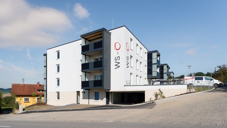 Moderne Wohnanlage mit Balkonen und Fahrzeugsicht im Hintergrund, sonniger Tag.