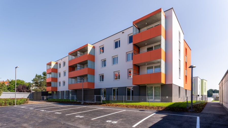 Modernes Wohngebäude mit orangefarbenen Balkonen, leere Parkplätze, sonniger Himmel.