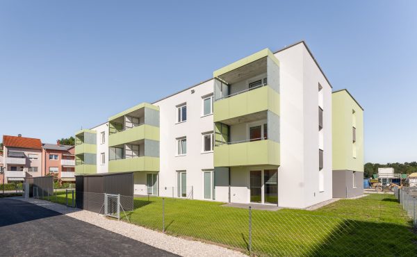 Mehrfamilienhaus mit grünen Balkonen und eingezäuntem Garten bei sonnigem Wetter.