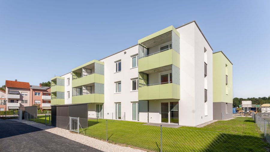 Mehrfamilienhaus mit grünen Balkonen und eingezäuntem Garten bei sonnigem Wetter.