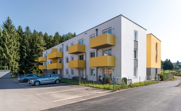 Mehrfamilienhaus mit gelben Balkonen und Autos im Vordergrund, umgeben von Bäumen.