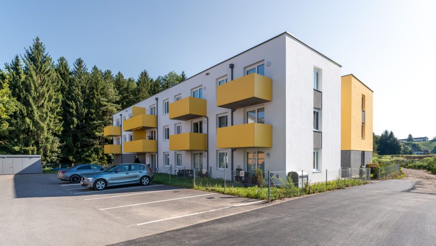 Mehrfamilienhaus mit gelben Balkonen und Autos im Vordergrund, umgeben von Bäumen.