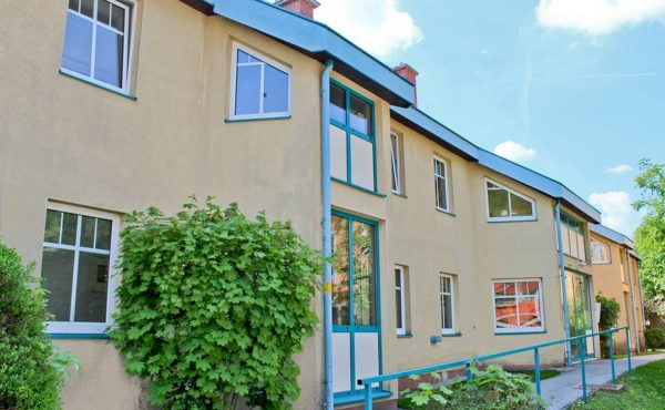 Gelbes Wohngebäude mit modernen Fenstern, umgeben von grüner Vegetation und klarem Himmel im Hintergrund.