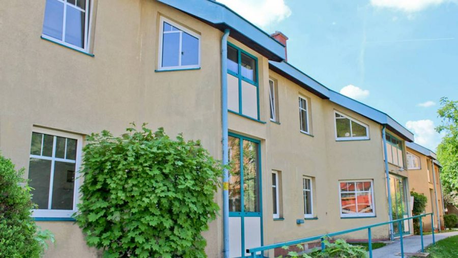 Gelbes Wohngebäude mit modernen Fenstern, umgeben von grüner Vegetation und klarem Himmel im Hintergrund.