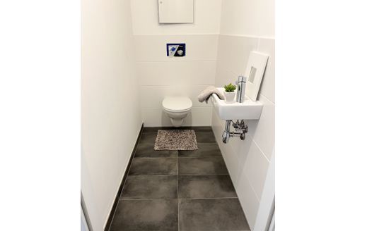 Modernes Badezimmer mit Toilette, kleinem Waschbecken und grauen Fliesen. Minimalistische Einrichtung und helle Wände.