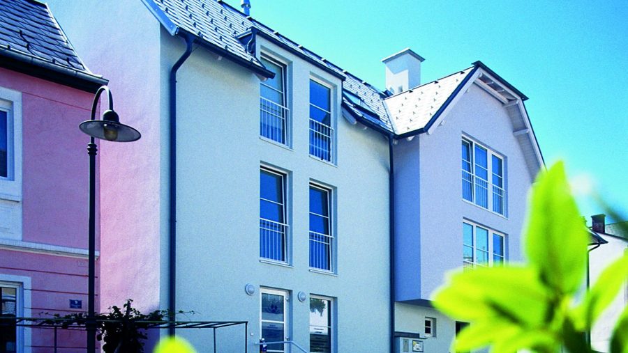 Mehrfarbiges Wohngebäude bei Sonnenschein, grüne Blätter im Vordergrund.