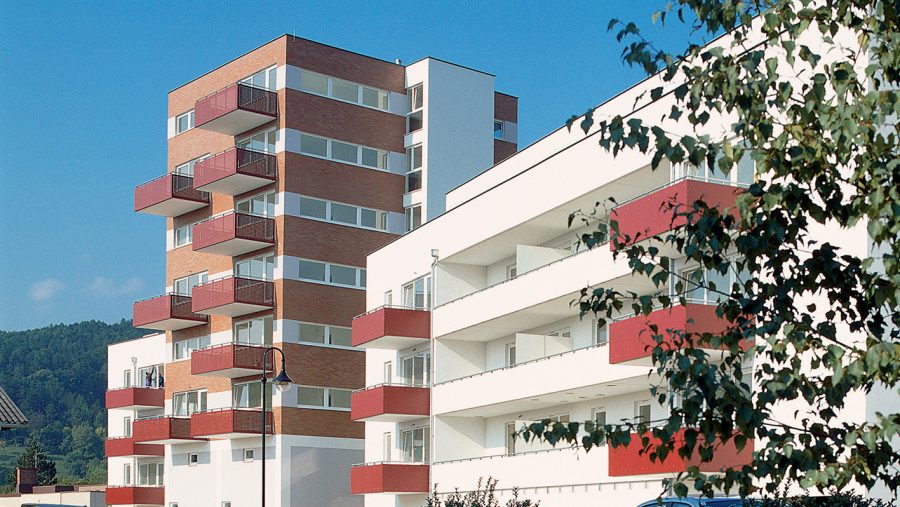 Mehrstöckige Wohngebäude mit roten Balkonen und Bäumen im Vordergrund an einem sonnigen Tag.