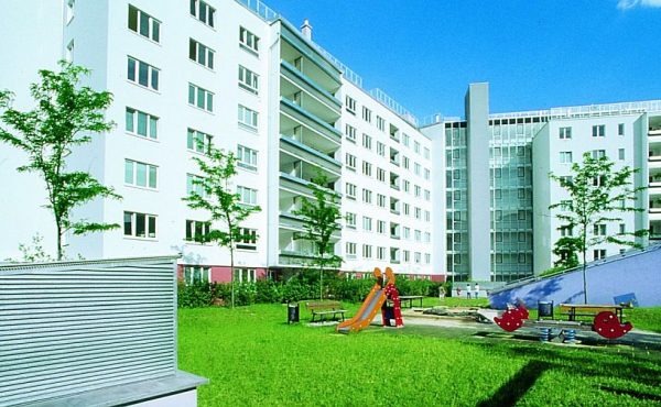 Mehrstöckiges Wohnhaus mit moderner Fassade und kleinem Spielplatz im Vordergrund, umgeben von grünem Rasen.