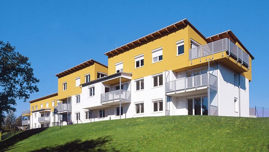 Moderne gelbe Wohnhäuser auf einem grünen Hügel vor klarem blauem Himmel.