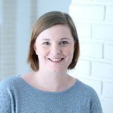 Frau mit braunen Haaren und blauem Pullover lächelt in heller Umgebung.