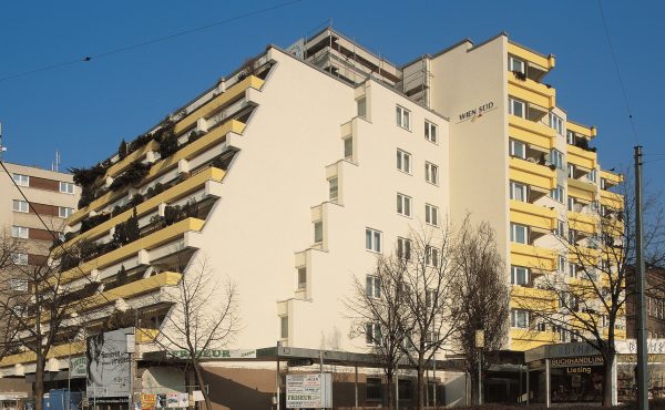 Mehrstöckiges Wohnhaus mit Balkonen und Geschäften, klarer blauer Himmel im Hintergrund.
