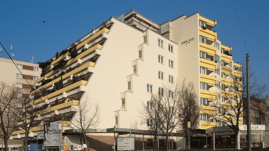 Mehrstöckiges Wohnhaus mit Balkonen und Geschäften, klarer blauer Himmel im Hintergrund.